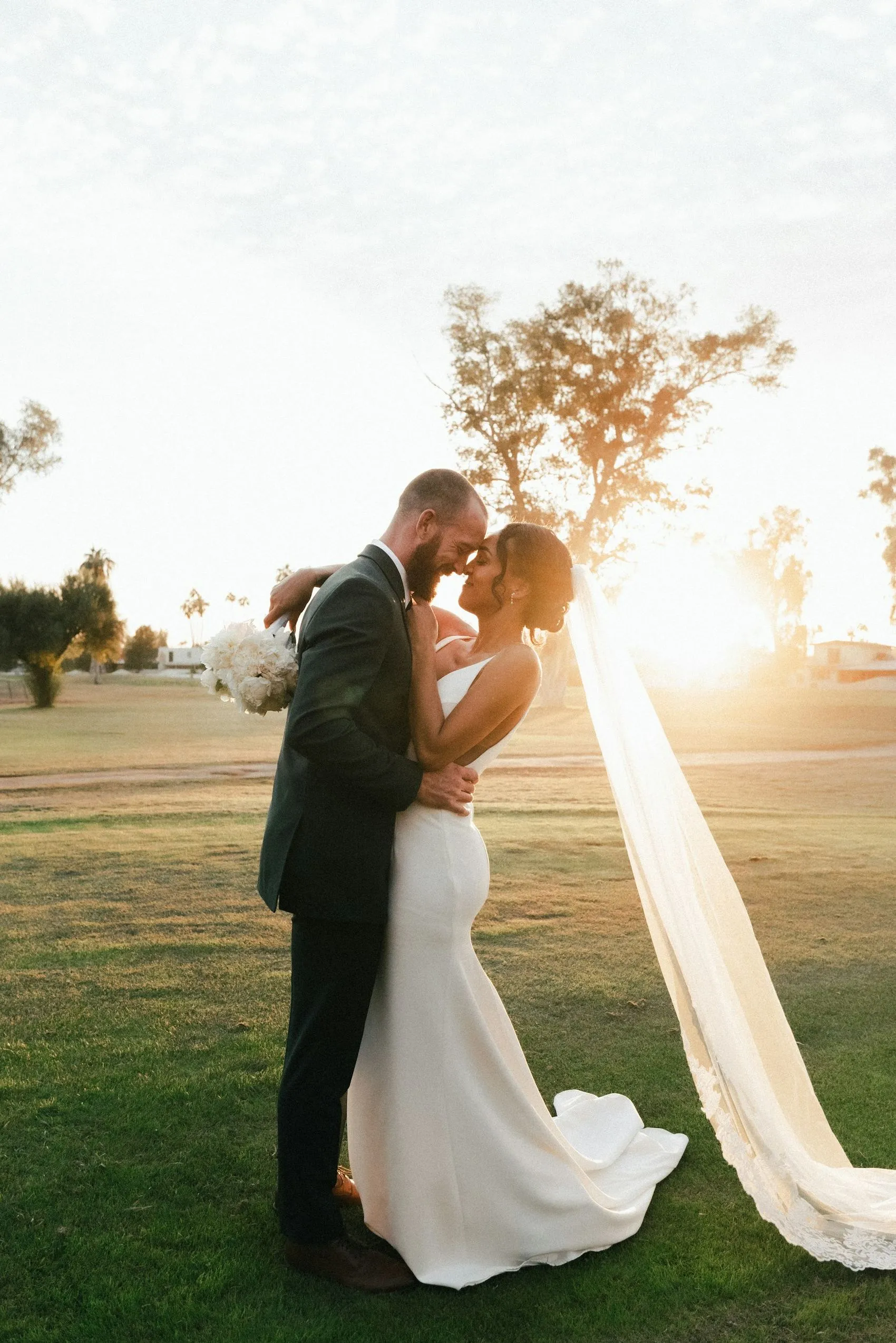 A couple mid-kiss in an open field at golden hour, the bride's veil caught in the wind